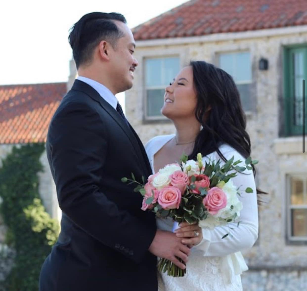 Bride and groom smile, gazing at each other in an outdoor setting, groom is wearing a black suit and white collared shirt, the bride is wearing long sleeve lace dress and holding a bouquet of fresh pink and white flowers.